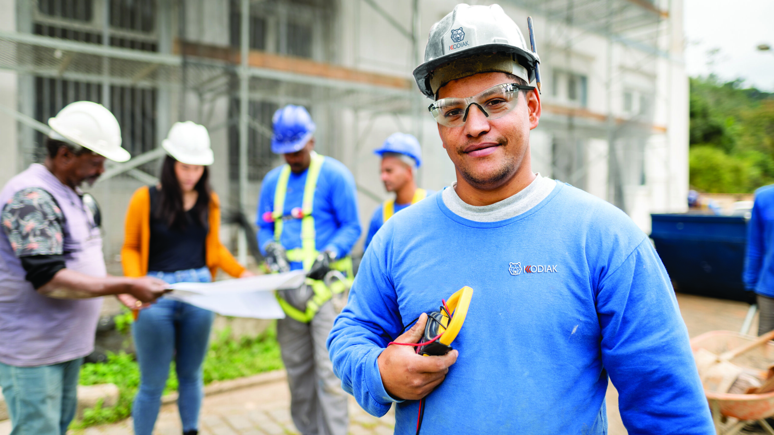 Portrait of a construction electrician holding a multimeter tool standing on a building site with people going over blueprints in the background